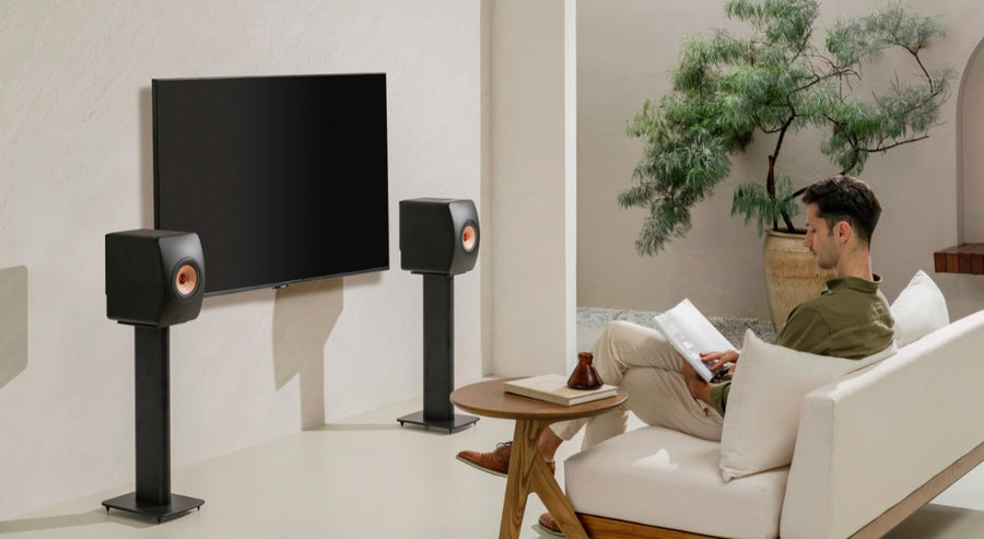 Man ontspant zich met een tijdschrift op een witte bank in een modern ingerichte woonkamer, met KEF-stereoluidsprekers op standaards naast een flatscreen-tv, een voorbeeld van een hoogwaardige home-entertainmentopstelling.