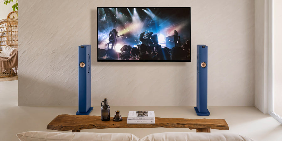 KEF LS60 Wireless speakers in een woonkamer met een tv ertussen waarop een live concert te zien is.