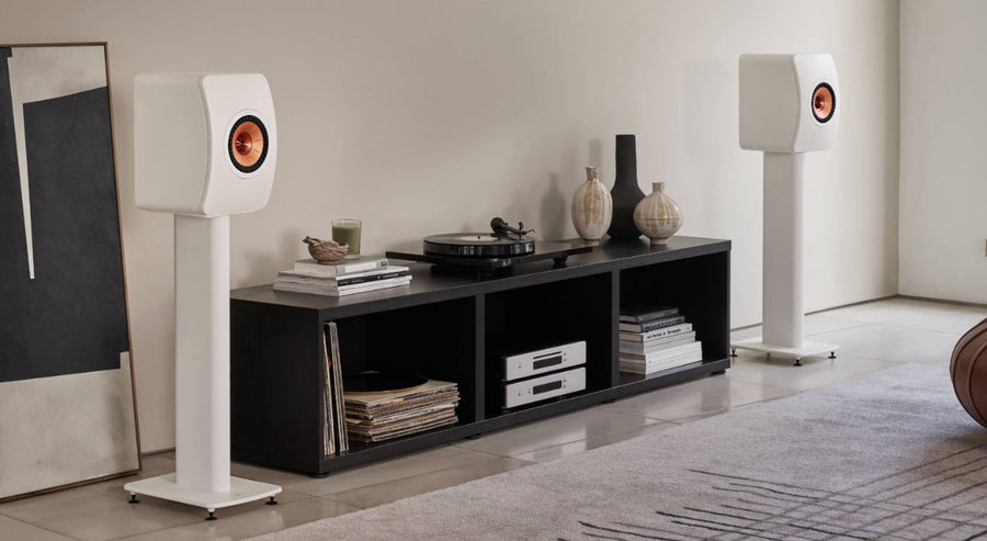 Moderne woonkamer met witte KEF‑boekenplankspeakers op stands, vinylplaten en een platenspeler op een laag mediameubel.