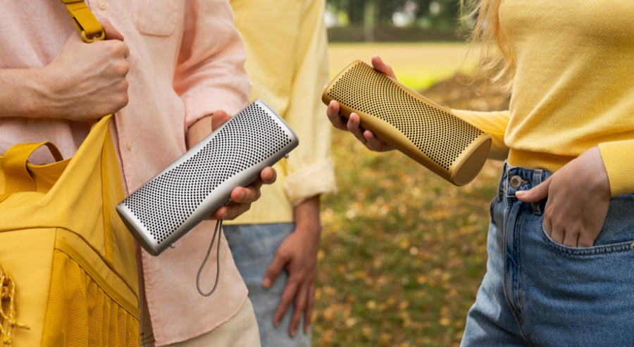 Twee mensen houden draagbare Bluetooth-speakers vast, één zilverkleurig en één goudkleurig, tijdens een informeel samenzijn in het park.